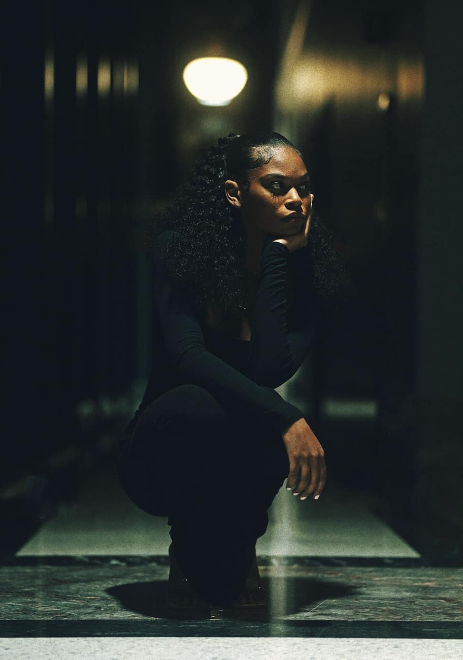 Modelo negra de preto agachada num corredor escuro, sob uma luz de cinema