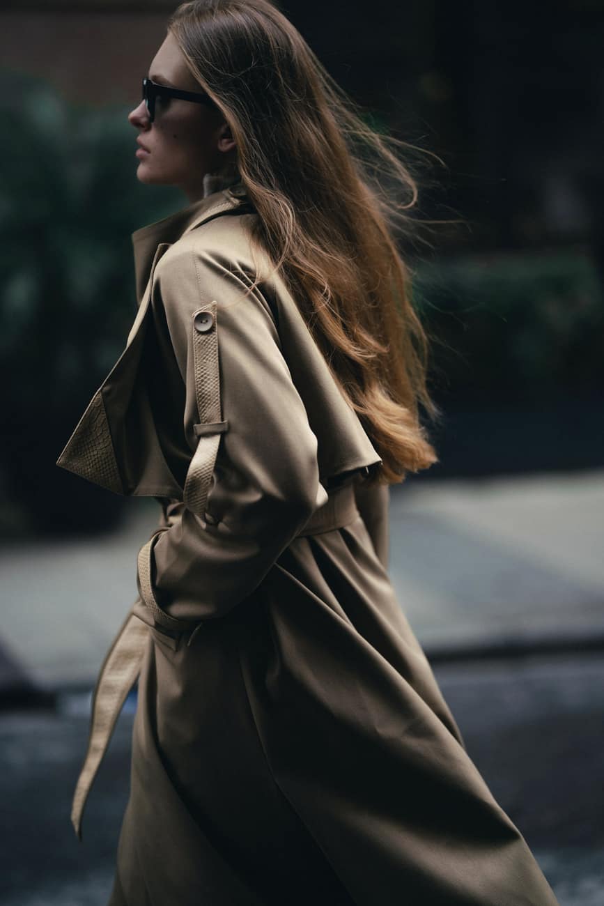 Mulher caminhando com trench coat cor de areia sobre fundo escuro