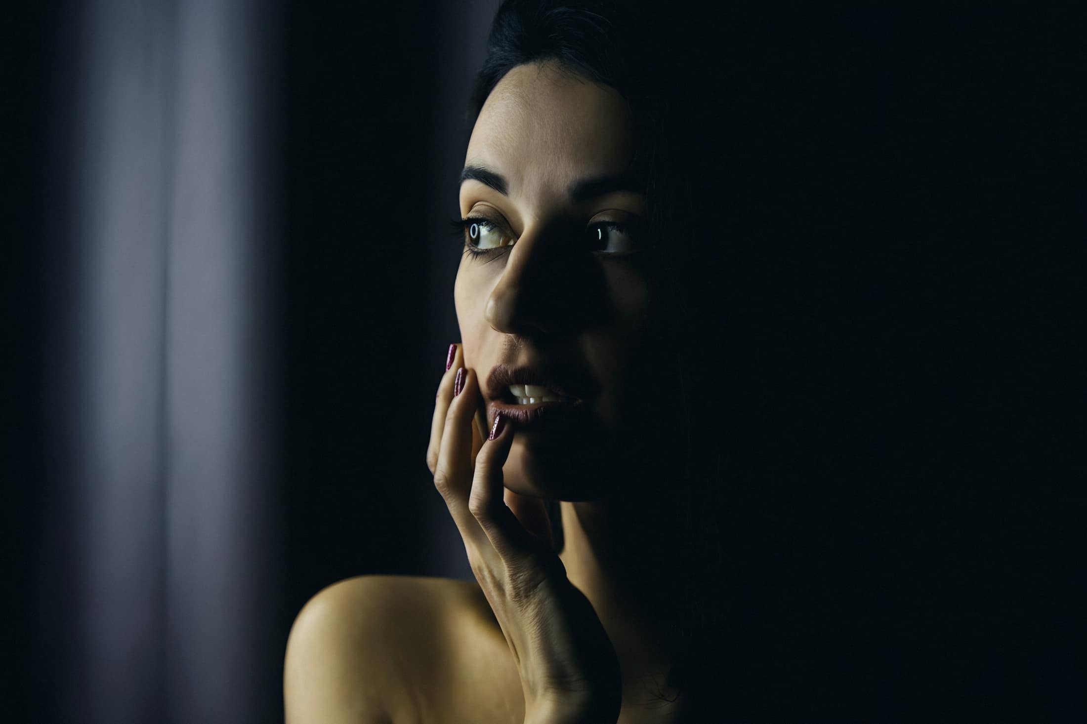 Close-up de mulher com a mão no rosto, em luz lateral sobre fundo escuro