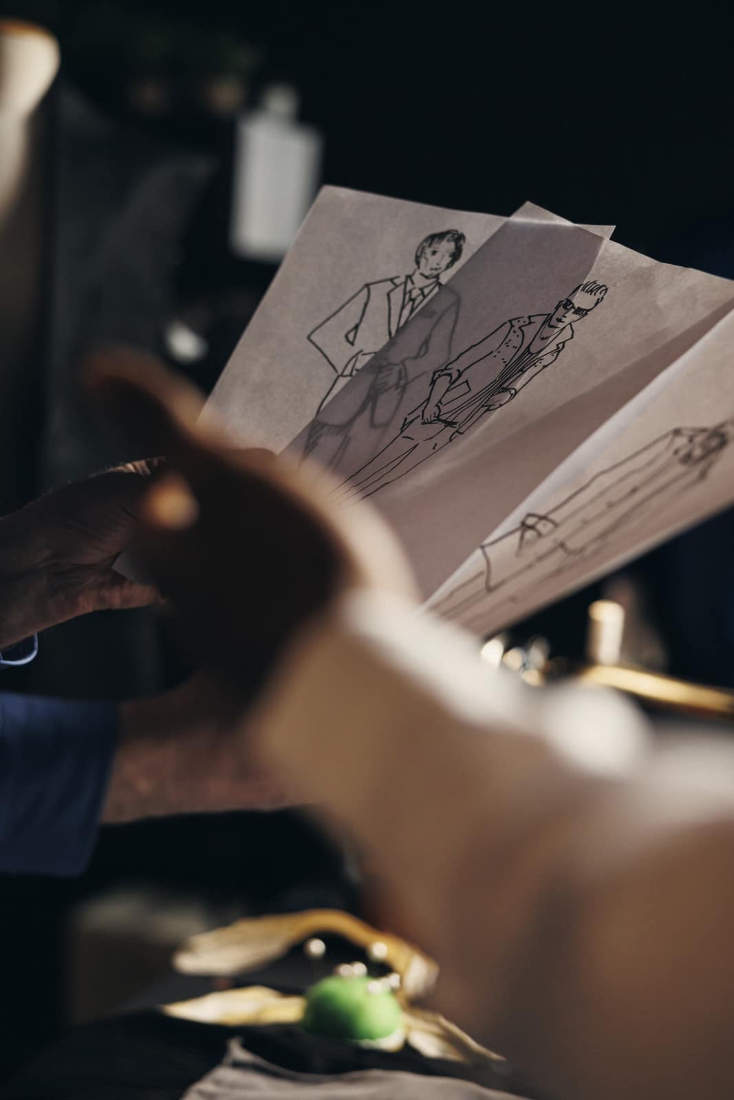 Mãos folheando croquis de alfaiataria na penumbra do ateliê